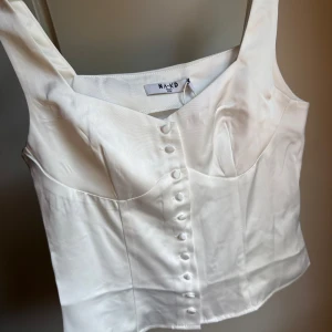Vit satin korsettopp från NA-KD - Supersnygg vit korsettopp i satin från NA-KD med breda axelband och dekorativa knappar framtill. Toppen har en figurnära passform och markerad byst, perfekt för att lyfta din look med en elegant touch.