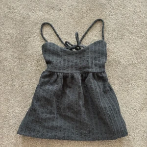 Svart babydoll topp med snörning - Supersöt svart babydoll topp med tunna axelband och snygg snörning i ryggen. Toppen har ett ribbat material. Perfekt för dig som gillar trendiga detaljer. 