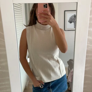 Beige stickad ärmlös polotopp - Säljer en beige stickad polotopp med hög krage och ärmlös design. Toppen har en clean look med diskreta sömdetaljer framtill och är perfekt att styla med jeans eller kjol. Materialet är mjukt och ger en snygg struktur.