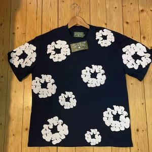 Mörkblå t-shirt från Denim Tears - Snygg mörkblå t-shirt från Denim Tears med stora vita blommiga tryck över hela tröjan. Klassisk passform med rund hals och korta ärmar. Perfekt statement-plagg för dig som vill sticka ut.