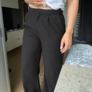 Svarta kostymbyxor - Säljer ett par svarta kostymbyxor med hög midja och vida ben. Byxorna har pressveck framtill, diskreta fickor i sidorna och en snygg, stilren look. Perfekta att styla med crop top eller skjorta för en clean vibe. Önskar du veta exakta mått? Tveka inte att höra av dig!
