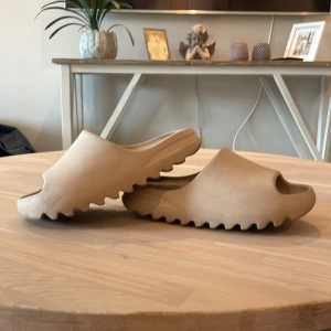 Adidas Yeezy Slides - Säljer ett par beige Adidas Yeezy Slides med chunky, sågtandad sula och öppen tå. Tillverkade i syntetmaterial för en mjuk och bekväm känsla. Perfekta för sommaren med sin minimalistiska design och neutrala färg som passar till allt. Ny pris 1300