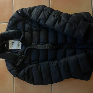 Svart pufferjacka från Tommy Hilfiger - Snygg svart pufferjacka från Tommy Hilfiger med klassisk quiltad design och hög krage. Jackan har dragkedja framtill och diskret logga på bröstet. Perfekt för kalla dagar när du vill hålla stilen och värmen.