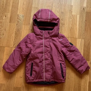 Vinröd vinterjacka från Lindberg, stl 110 - Snygg vinröd vinterjacka från Lindberg med huva och reflexdetaljer. Jackan har dragkedja framtill, två fickor med svarta dragkedjor och är fodrad för extra värme. Materialet är nylon med polyesterfyllning, perfekt för kalla dagar.