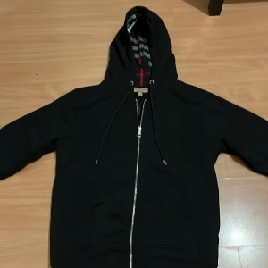Svart hoodie från Burberry med dragkedja - Svart hoodie från Burberry med hel dragkedja och klassiskt rutigt foder i huvan. Tröjan har snörning i huvan och diskret broderad logga på bröstet. Perfekt för dig som gillar stilrena och exklusiva plagg med snygga detaljer. Pris kan diskuteras vid snabb och smidig affär😊😊