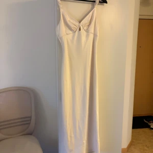 Champagnefärgad långklänning från Adoore - Supersnygg långklänning från Adoore i champagnefärgad satin. Klänningen har smala axelband med rynkade detaljer, v-ringning och en liten cut-out framtill. Sval och elegant passform som ger en lyxig känsla. Perfekt för dig som vill sticka ut med stil.