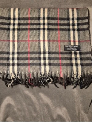 Rutig halsduk från Burberry i ull - Klassisk halsduk från Burberry i 100% lammull med ikoniskt rutigt mönster i grått, svart, vitt och röda detaljer. Mjuk och varm med fransar i kanterna och diskret Burberry-märke. Perfekt accessoar för att lyfta din outfit.