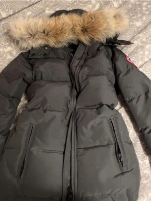 Mörkgrå Canada Goose jacka  - Säljer en mörkgrå dunjacka från Canada Goose med avtagbar huva och snygg pälsdetalj. Jackan har långa ärmar, två stora fickor med dragkedja och klassisk patch på ärmen. Perfekt för kalla vinterdagar och riktigt varm. Såklart är pälsen äkta! Fler bilder kan skickas privat, bara att kontakta mig😊