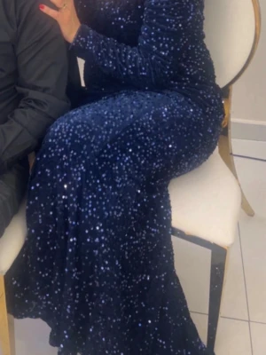 Marinblå glittrig långklänning - Säljer en marinblå långklänning täckt av glittriga paljetter. Klänningen har lång ärm och figurnära passform som ger en elegant siluett. Perfekt för dig som vill sticka ut på bal eller fest. Materialet känns lyxigt och skimrar snyggt i ljuset.