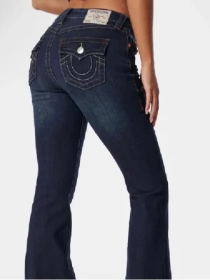 Mörkblå bootcut jeans från True Religion - Säljer ett par mörkblå jeans från True Religion med bootcut passform i Becca. Jeansen har kontrastsömmar, ikoniska fickdetaljer med lock och knapp bak samt klassisk femficksdesign. Tillverkade i slitstarkt denim med snygg tvätt och låg midja.