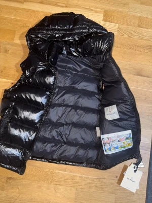 Svart dunväst från Moncler - Snygg svart dunväst från Moncler med glansig finish och huva. Västen har två dragkedjeförsedda fickor framtill, quiltad design och klassisk Moncler-logga på bröstet. Perfekt lager på lager-plagg för kyligare dagar. Märkeslappar och serietikett finns kvar. NFC TAGGAD, HELT NY