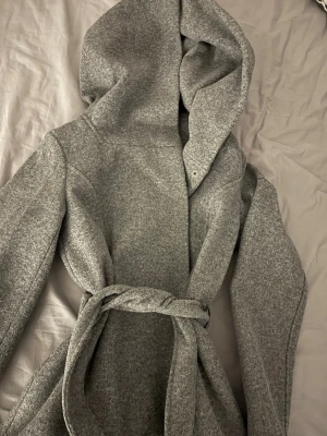 Grå kappa med huva och bälte - Säljer en stilren grå kappa med huva och knytbälte i midjan. Jackan har lång ärm och är tillverkad i ett mjukt ull-liknande material. Perfekt för kyliga dagar när du vill vara både varm och snygg.