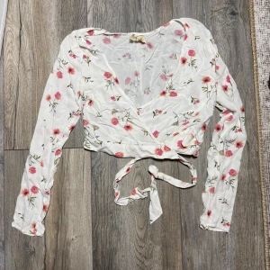 Blommig omlottopp från Hollister XS - Supersöt omlottopp från Hollister i vitt med blommigt mönster i rosa och beige toner. Toppen har långa ärmar, v-ringning och knytband i midjan för en snygg passform. Perfekt till jeans eller kjol för en fräsch look.