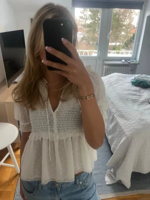 Vit blus med volang och knyt - Supersöt vit blus med korta puffärmar, volangdetaljer och knyt framtill. Blusen har smock över bysten och är tillverkad i ett lätt, transparent tyg med diskreta prickar. Perfekt till jeans för en somrig look.