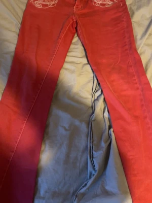 Röda jeans med broderi och patch - Snygga röda jeans med vita broderade detaljer på bakfickorna och längs linningen. Byxorna har en rak passform och ett coolt tygmärke med krona och vingar bak i midjan. Perfekta för dig som vill sticka ut lite extra. Klassisk femficksmodell i jeansmaterial.