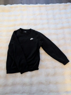Svart sweatshirt från Nike - Klassisk svart sweatshirt från Nike med vit logga på bröstet. Tröjan har rund halsringning, ribbade muddar och är långärmad. Perfekt för en chill och sportig look.