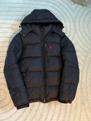 Svart pufferjacka från Polo Ralph Lauren - Svart pufferjacka från Polo Ralph Lauren med huva och röd logga. Jätte bra skick ser helt orörd ut vilket den nästan är! Skriv för fler frågor.