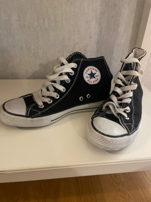 Svarta Converse All Star high tops - Säljer ett par klassiska svarta Converse All Star high tops med vit sula och vita snören. Skorna har rund tå, platt sula och tydliga vita sömmar. Märket syns på sidan och tungan. Materialet är bomullstyg och gummisula. Perfekt för dig som gillar ikoniska sneakers.