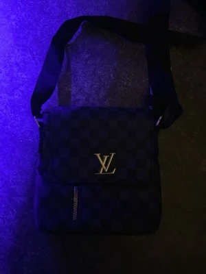 Svart rutig axelväska - Snygg svart axelväska  med klassiskt rutigt mönster och silverfärgad loggan på framsidan. Väskan har justerbar axelrem, dragkedja framtill och magnetlås. Materialet är skinn och polyester, perfekt för dig som gillar stilrena accessoarer.