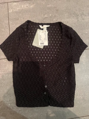 Svart blus med hålmönster från H&M - Kortärmad svart blus från H&M i storlek XS. Toppen har ett snyggt hålmönster, v-ringning och knappar framtill. Materialet är en mix av bomull och polyester, vilket gör den både mjuk och luftig. Perfekt till jeans eller kjol för en trendig look.