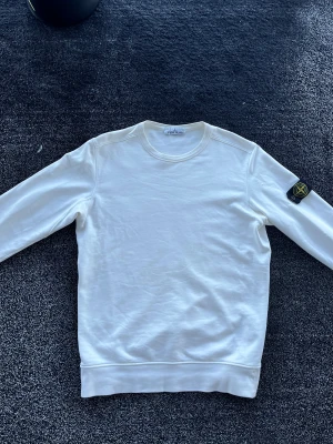 Stone Island sweatshirt - Säljer en clean vit sweatshirt från Stone Island med klassisk patch på vänster ärm. Tröjan har rund halsringning och är tillverkad i mjuk bomull. Perfekt för dig som gillar stilrena plagg med streetwear-vibe. Storlek L, skick 7/10 har några små defekter vilket syns på den tredje bilden annars bra skick, äkta. Pris går att diskutera✅