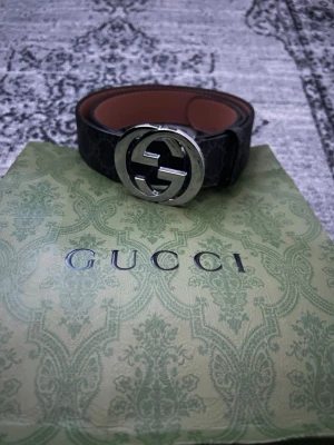Gucci svart bälte  - Säljer ett svart bälte från Gucci med det klassiska GG-mönstret och en stor, rund silverfärgad GG-logga som spänne. Bältet har en lyxig känsla och insidan är brun. Perfekt accessoar för att lyfta vilken outfit som helst.