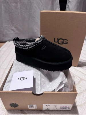 Svarta UGG w tazz  - UGG slip-ons i svart mocka med fluffigt foder och platåsula. Skorna har dekorativt mönster i vitt runt öppningen och UGG-logga på sidan. Perfekta för kalla dagar när du vill ha både stil och komfort.