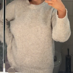 Beige stickad tröja med rund hals - Mysig beige stickad tröja med rund hals och långa ärmar. Tröjan har en avslappnad passform och ribbade muddar vid hals, ärmslut och nederkant. Perfekt för kyliga dagar och enkel att matcha med jeans eller kjol.