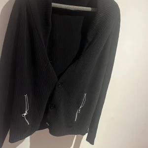 Svart ribbad kofta med dragkedjor - Säljer en svart ribbad kofta med knappar framtill och två coola dragkedjor vid fickorna. Koftan har lång ärm och en bred krage som ger en avslappnad vibe. Perfekt för lager på lager och funkar till det mesta i garderoben.