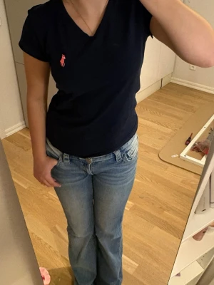 Mörkblå t-shirt från Polo Ralph Lauren - Snygg mörkblå t-shirt från Polo Ralph Lauren med rosa logga broderad på bröstet. Modellen är kortärmad med v-ringning och har en normal passform. Tillverkad i 100% bomull för en skön känsla. STORLEKEN SÄGER M men passar S