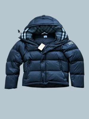 Svart pufferjacka från Burberry - Säljer en svart pufferjacka från Burberry med avtagbara ärmar så den kan bäras som väst. Jackan har huva med klassiskt rutigt foder, dragkedja och knappar framtill samt två fickor med dragkedja. Perfekt för kalla dagar och riktigt snygg streetstil.