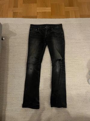 Tornado Mart Jeans - Säljer ett par svarta slim/bootcut jeans med coola slitningar och hål på benen. Jeansen har en smal passform och klassisk femficksdesign. Färgen är svart med lite tvättade detaljer för en snygg, sliten look. Perfekta för dig som gillar en edgy stil.