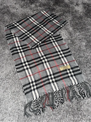 Rutig cashmerehalsduk från Burberry - Säljer en klassisk rutig halsduk från Burberry i 100% kashmir. Halsduken har grå, svarta, vita och röda rutor och avslutas med tvinnade fransar. Perfekt accessoar för att lyfta din outfit med en tidlös brittisk touch.