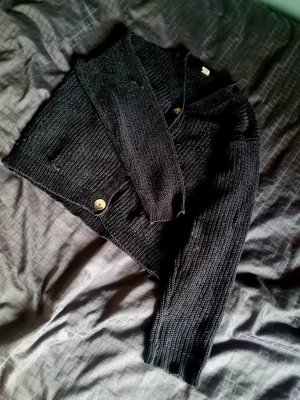 Svart stickad kofta från Only Young - Mysig svart stickad kofta från Only Young med grov ribbad struktur och stora, kontrasterande knappar framtill. Koftan har lång ärm och en loose passform som ger en chill vibe. Perfekt att slänga på sig när det är lite kyligt ute.