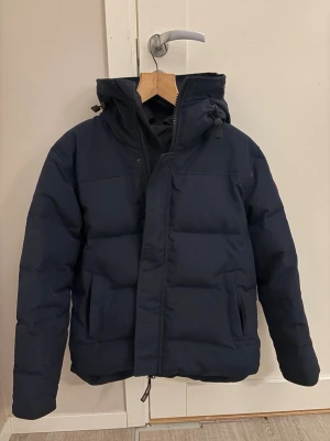 Mörkblå dunjacka från Canada Goose - Säljer en mörkblå dunjacka från Canada Goose med huva och dragkedja. Jackan har två sidofickor och klassisk patch på ärmen. Perfekt för kalla vinterdagar och har en stilren, enkel design.