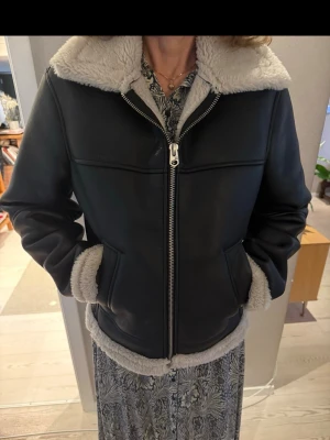 Svart teddyjacka från Zara med fuskpäls - Säljer en svart teddyjacka från Zara i storlek S. Jackan har mjukt beige fuskpälsfoder, stor krage och dragkedja framtill. Fodret syns även vid ärmslut och nederkant. Perfekt för kalla dagar. 