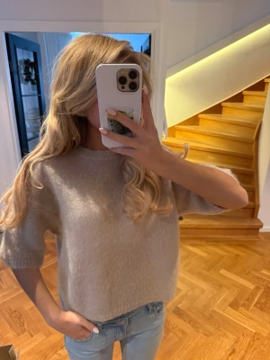Beige stickad tröja med kort ärm - Säljer en beige stickad tröja med korta ärmar och rund hals. Tröjan har en mjuk och fluffig känsla, är lite croppad och har en enkel, stilren design. Perfekt att matcha med jeans för en chill look.
