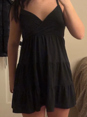 Volang Klänning - Svart kortklänning från Hollister i storlek S. Klänningen har omlottdesign vid bysten, smala axelband och volangdetaljer i kjolen. Tillverkad i mjuk bomull som känns skön mot huden. Perfekt för dig som gillar enkel och stilren look.