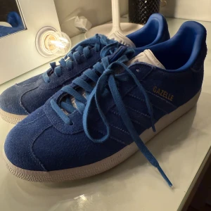 Adidas Gazelle blå sneakers - Säljer ett par Adidas Gazelle sneakers i blå mocka med vita detaljer och guldtext på sidan. Skorna har klassisk snörning, rund tå och platt sula. Insidan är blå och tungan är vit med Adidas-logga. Perfekta för dig som gillar retrostil och vill sticka ut.