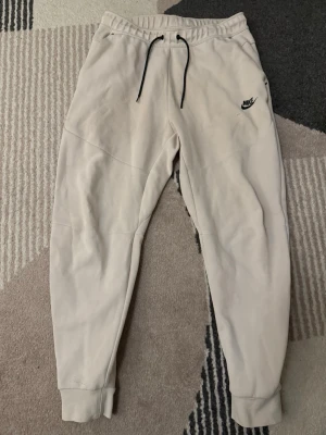 Beige mjukisbyxor Nike tech - Säljer ett par beige mjukisbyxor från Nike med svart logga på vänster ben. Byxorna har resår i midjan med svart snörning och muddar vid bensluten. Perfekta för chill eller träning, riktigt sköna och stilrena.