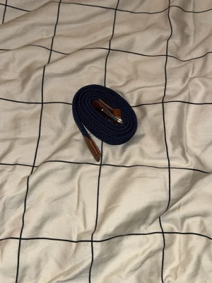 Mörkblå grisch bälte med läderdetaljer - (HELT NYTT)Snyggt mörkblått flätat bälte med bruna läderdetaljer vid spännet och ändarna. Bältet har ett klassiskt metallspänne och är flexibelt i materialet, vilket gör det enkelt att justera. Perfekt för att ge outfiten en stilren touch.