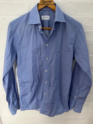 Blå prickig skjorta från Eton - Snygg blå skjorta från Eton i slim fit med små vita prickar. Klassisk krage, långa ärmar och vita knappar framtill. Tillverkad i mjuk bomull som känns skön mot huden. Perfekt för dig som gillar stilrena detaljer och en modern passform.