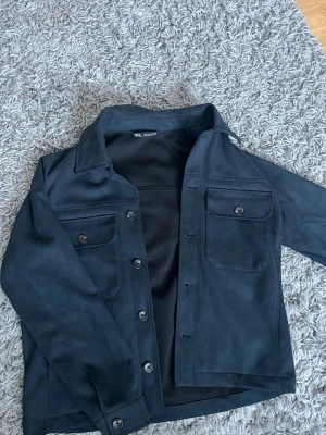 Svart mocka overshirt från Zara - Säljer en svart overshirt från Zara i mjuk mocka-look. Jackan har klassisk krage, två stora bröstfickor med knappar och stängs med knappar framtill. Perfekt att slänga över en hoodie eller t-shirt för en clean och stilren vibe.