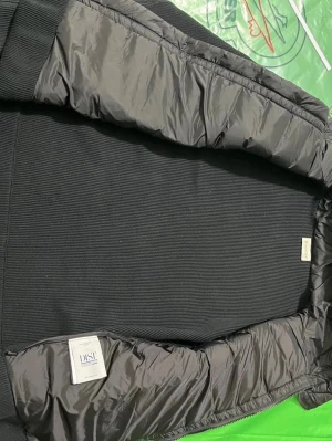 Svart cardigan  från Moncler - Säljer en svart cardigan från Moncler med stickade ärmar och huva. Jackan har dragkedja framtill, fickor med dragkedja och en Moncler-logga på ärmen. Materialmix av polyester och stickad bomull ger en modern look. Perfekt för kyliga dagar.