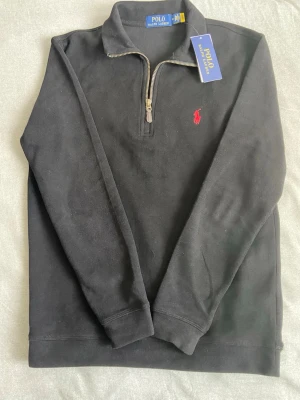 Svart half zip tröja från Polo Ralph Lauren - Svart långärmad tröja från Polo Ralph Lauren med half zip och klassisk röd broderad logga på bröstet. Tröjan har en ståkrage och är gjord i mjuk bomull, perfekt för chill höst- och vinterdagar. Snygg och enkel att matcha med jeans eller chinos. Storleken är M men skulle säga att den passar mer som en S.