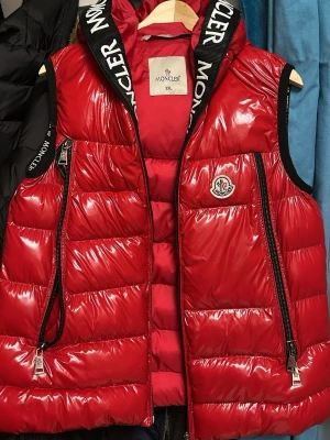 Röd dunväst från Moncler - • Märke: Moncler • Typ: Väst / puffer-väst • Färg: Röd • Material: Blank nylon • Fyllning: Dun • Storlek: XXL • Passform: Passar perfekt även för dig som vanligtvis har S, M eller L (snygg oversize-look) • Huva • Dragkedja framtill • Fickor med dragkedja • Moncler-logga på bröstet  Västen är i mycket fint skick, inga fel eller skador. Säljer endast för att jag vill köpa en ny jacka.