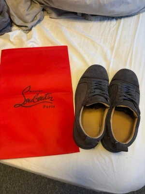 Mörkgrå mocka sneakers från Louboutin - Säljer ett par stilrena mörkgrå sneakers från Christian Louboutin i mjuk mocka. Skorna har rund tå, platt sula och klassisk snörning. Insidan är beige och de kommer med original röd dustbag. Perfekta för dig som gillar lyxiga detaljer och minimalistisk design.