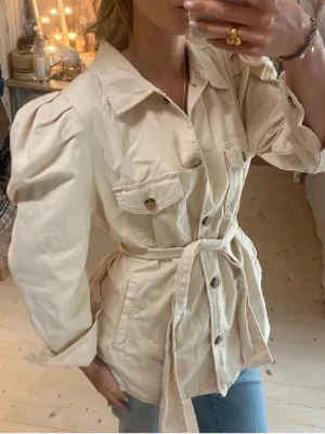 Beige overshirt med bälte  - Snygg beige overshirt med puffade axlar, stora bröstfickor och knappar framtill. Jackan har ett matchande bälte i midjan och är tillverkad i mjuk bomull. Perfekt att styla med jeans för en chill look.