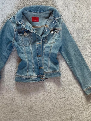Blå jeansjacka från Cross Jeans - Säljer en klassisk blå jeansjacka från Cross Jeans i storlek S. Jackan har långa ärmar, framfickor med knappar och snygga sömdetaljer. Perfekt att slänga över en t-shirt eller hoodie för en chill look.