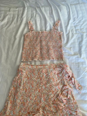 Blommig linnetopp och kjol från KappAhl - Supersöt linnetopp med kjol från KappAhl i storlek 170. Toppen har ett smockat tyg med volangkant och breda axelband. Mönstret är blommigt i pastelliga toner av rosa, orange, beige och lite grönt. Perfekt för sommarens alla dagar!